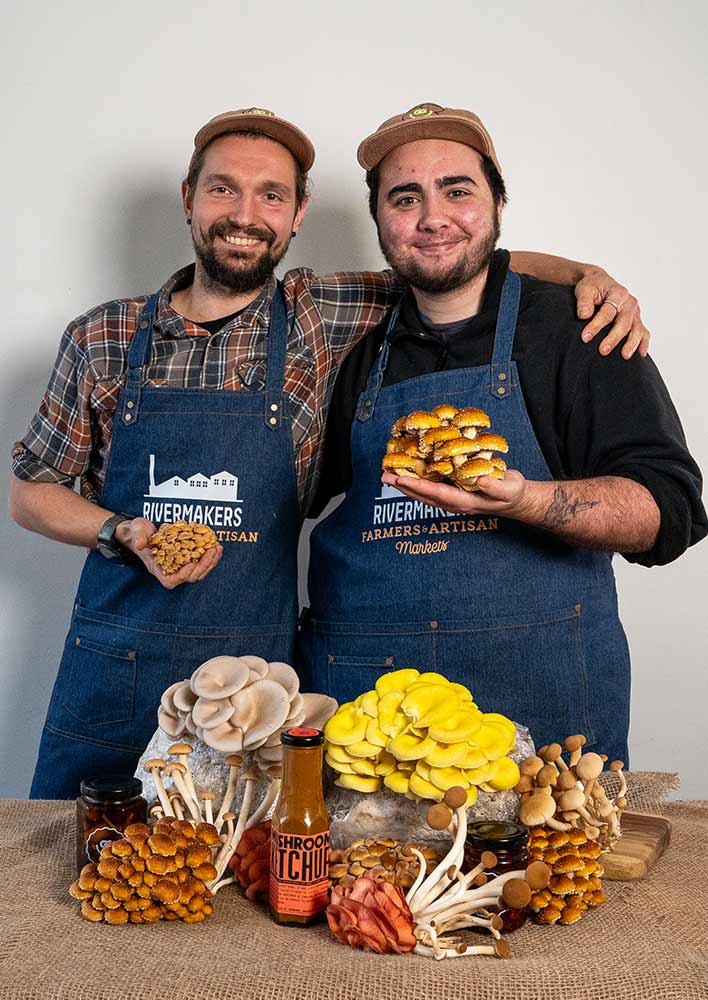 Two men wearing aprons looking at the camera holding mushrooms